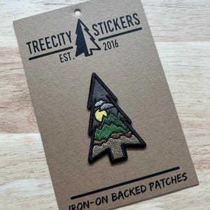 May include: An iron-on patch in the shape of a tree, featuring a mountain scene with a yellow moon. The patch is on a brown card with the text "TREECITY STICKERS EST. 2016" and "IRON-ON BACKED PATCHES".