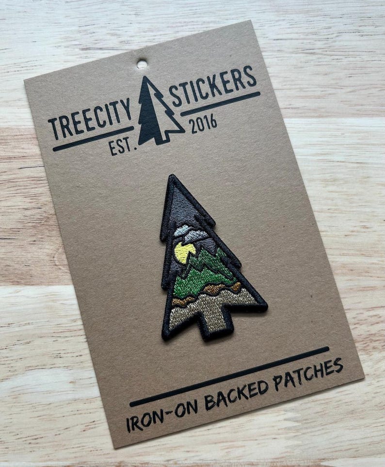 May include: A tree-shaped iron-on patch with a mountain scene and a yellow moon. The patch is on a brown card with the text "TREECITY STICKERS EST. 2016" and "IRON-ON BACKED PATCHES".