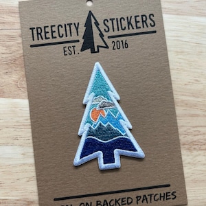May include: A colorful iron-on patch in the shape of a stylized evergreen tree, featuring a mountain scene with an orange sun. The patch is on a brown card with the text "TREECITY STICKERS EST. 2016" and "IRON-ON BACKED PATCHES".
