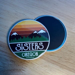May include: A round magnet featuring a graphic of snow-capped mountains above a green forest and a sunset sky with orange and yellow stripes. The text "SISTERS" appears in white letters on a dark band, with the word "OREGON" printed below in white.