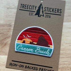 May include: A semi-circular embroidered patch featuring a sunset landscape of Cannon Beach with a red sky, orange sun, and dark rock formations. The patch has a teal base with the text Cannon Beach in white script and a white border.
