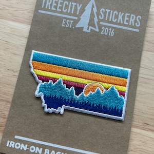 May include: An iron-on patch featuring a colorful landscape design of the state of Montana. The patch has a blue, orange, yellow, and pink striped sky, with a mountain range and a setting sun. The patch is on a brown card with the text "TREECITY STICKERS" and "IRON-ON BACKED PATCHES".