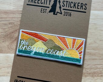 May include: Embroidered patch depicting a sunset over mountains with the text "the oregon coast." The patch is rectangular with a white border and is attached to a brown card with the text "Treecity Stickers" and "Iron-on backed patches."