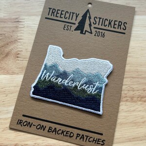 May include: An embroidered patch shaped like the state of Oregon, featuring a mountain landscape in shades of blue, green, and white with the word Wanderlust stitched in white. It is attached to a brown card labeled Treecity Stickers Est. 2016.