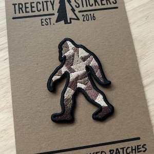 May include: Embroidered patch featuring a Bigfoot design with a black outline. The Bigfoot is filled with a patchwork of brown, tan, and cream colors. The patch is on a brown card with the text "TREECITY STICKERS EST. 2016".