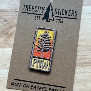May include: A rectangular iron-on patch featuring an embroidered design of a dark grey pine tree against a yellow and orange gradient background. The letters PNW are stitched in dark grey at the bottom. The patch is mounted on a brown display card.