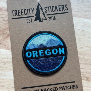 May include: A circular embroidered patch featuring a mountain landscape in shades of light and dark blue, with the word OREGON in bright blue text across a black horizontal band. The patch is attached to a brown card labeled TREECITY STICKERS.