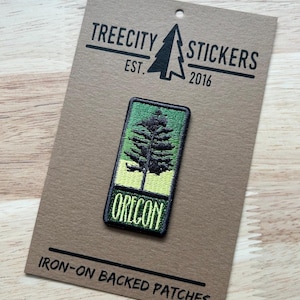 May include: A rectangular iron-on patch featuring a green and black tree design with the word "OREGON" in green lettering. The patch is on a brown card with the text "TREECITY STICKERS EST. 2016" and "IRON-ON BACKED PATCHES".