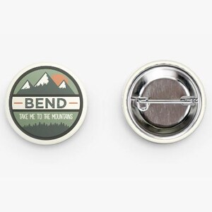 May include: A circular pin-back button featuring a graphic of mountains and a forest in shades of green, white, and orange. The text BEND is centered in the middle, with the phrase TAKE ME TO THE MOUNTAINS printed below it. The back shows a metal pin.