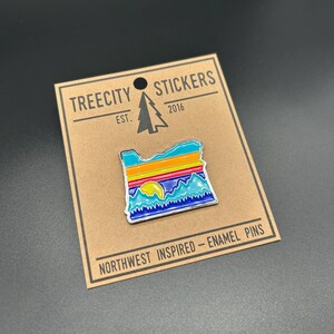 May include: A metal enamel pin shaped like the state of Oregon, featuring a colorful landscape with a yellow sun, blue mountains, and horizontal stripes of orange, yellow, and teal. It is attached to a brown card labeled TREECITY STICKERS.