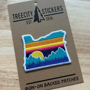 May include: An embroidered iron-on patch shaped like the state of Oregon, featuring a sunset landscape with horizontal stripes of teal, orange, yellow, and pink above a dark blue mountain range and a yellow sun on a brown branded display card.
