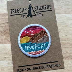 May include: A round embroidered patch featuring a coastal landscape with the word "NEWPORT" in teal. The patch has a sunset sky, brown hills, and teal water. The backing card reads "TREECITY STICKERS EST. 2016" and "IRON-ON BACKED PATCHES."