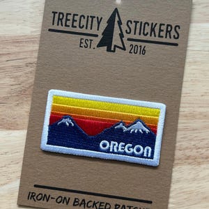 May include: A rectangular iron-on embroidered patch featuring a landscape of dark blue mountains with white snow-capped peaks under a sunset sky of yellow, orange, and red horizontal stripes. The word OREGON is stitched in white at the bottom.