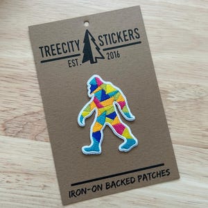 May include: An embroidered patch featuring a geometric, multi-colored Bigfoot silhouette in shades of pink, yellow, blue, and green. The patch is attached to a brown cardboard display card printed with the text TREECITY STICKERS EST. 2016 and IRON-ON BACKED PATCHES.