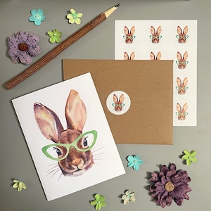 May include: A white card with a watercolor illustration of a brown rabbit wearing green glasses. The card is on a brown envelope with a sheet of stickers featuring rabbits wearing glasses.
