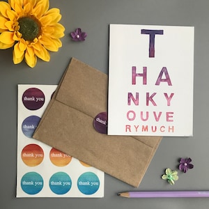 May include: A watercolor thank you card with the words "THANK YOU VERY MUCH" in purple and pink. The card is next to a brown envelope, round stickers with "thank you" text, a yellow sunflower, and a purple pencil.