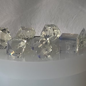 May include: A set of seven clear glass dice with etched numbers. The dice are in various shapes, including a pyramid, a cube, and a 20-sided die.