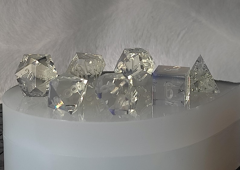 May include: A set of seven clear glass dice with etched numbers. The dice are in various shapes, including a pyramid, a cube, and a 20-sided die.