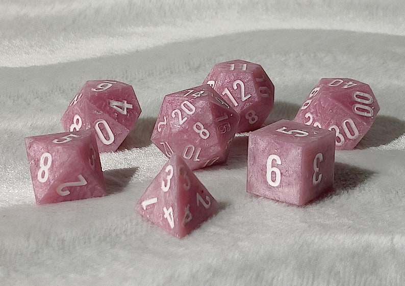May include: A set of pink dice with white numbers. The dice are in various shapes, including a d4, d6, d8, d10, d12, and d20.