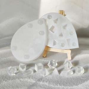May include: Two white silicone molds for making resin dice. The molds are shaped like a teardrop and a heart. There are several clear resin dice on a white fuzzy surface.