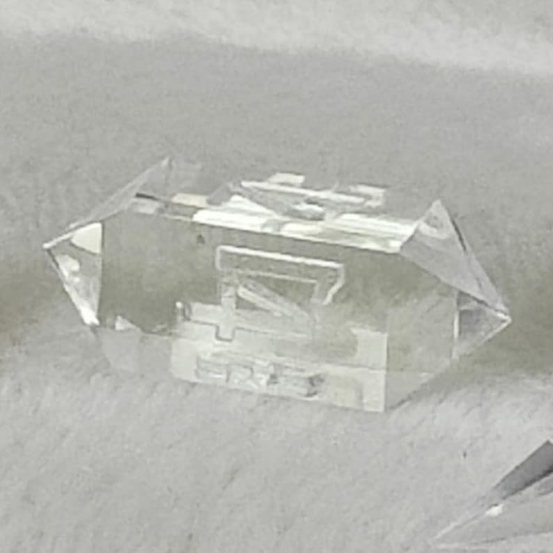 May include: A clear, faceted crystal with a geometric shape. The crystal is shaped like a long, narrow pyramid with a pointed top and a flat base. The crystal is translucent and has a slight shimmer.