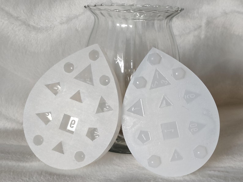 May include: Two white silicone molds shaped like teardrops. Each mold has a different set of shapes for making dice. One mold has a d4, d6, d8, d10, d12, and d20. The other mold has a d4, d6, d8, d10, d12, and d20.