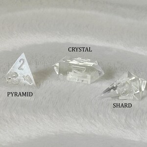 May include: Three clear glass dice with different shapes: a pyramid, a crystal, and a shard. The pyramid die has the number 2 on it. The crystal die has the number 7 on it. The shard die has the number 5 on it.