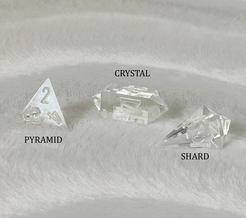 May include: Three clear glass dice with different shapes: a pyramid, a crystal, and a shard. The pyramid die has the number 2 on it. The crystal die has the number 7 on it. The shard die has the number 5 on it.