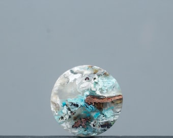 May include: A round gemstone with a unique pattern. The stone features a mix of colors, including white, blue, brown, and hints of black. The overall design resembles a landscape or abstract art piece. The gemstone is set against a gray background.