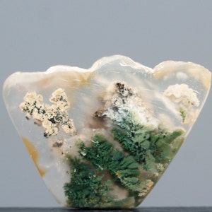 Może przedstawiać: Przejrzysty, biały agat z zielonymi i brązowymi wrostkami mchu. Agat ma kształt wachlarza i gładką, polerowaną powierzchnię.