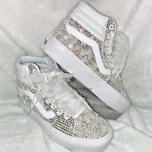 Könnte beinhalten: Weiße High-Top-Sneaker, die mit Strasssteinen und Perlen besetzt sind und eine weiße Tüllschleife haben. Die Schuhe befinden sich auf einer weißen, flauschigen Oberfläche.