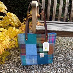 May include: A patchwork tote bag with brown leather handles. The bag is made of various blue, green, and brown plaid and solid fabric squares. A white tag hangs from the side. The bag is sitting on a bed of small rocks.
