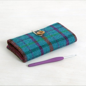 May include: A teal and purple plaid crochet hook case with a wooden button closure. A purple crochet hook rests beside the case. The case is made of a textured fabric.
