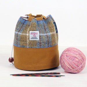 May include: A Harris Tweed drawstring yarn bag with a blue, brown, and white plaid upper and a tan base. A ball of pink and white yarn and two pairs of knitting needles are next to the bag. The bag has a label that says "Harris Tweed".