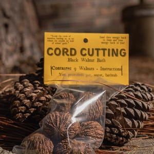 Puede incluir: Un paquete de nueve nueces negras para un baño de nueces negras Cord Cutting, con instrucciones en una tarjeta amarilla. La imagen también muestra piñas y una superficie de madera.