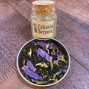 May include: A small glass jar with a cork lid labeled "Smell Me" sits above a tin filled with loose leaf tea. The tea is a mix of dark green leaves and vibrant purple and yellow flower petals. The jar has a label that reads "Celestial Serpent".
