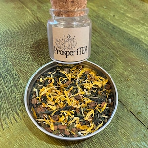 May include: A glass jar with a cork lid filled with loose leaf tea. The jar has a brown paper label with the text "ProsperiTEA" in black lettering. The tea is a blend of green tea, dried fruit, and herbs.