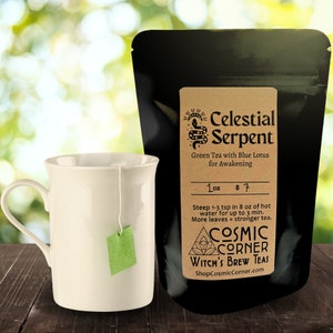 Op de afbeelding: Een witte keramische mok met een groen theezakje naast een zwarte zak met het opschrift "Celestial Serpent" met "Green Tea with Blue Lotus for Awakening". De zak zegt ook "1 oz".