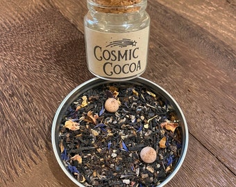 May include: A glass jar with a cork lid, labelled "Cosmic Cocoa", sits above a small tin filled with loose tea. The tea blend includes dried flowers and two round, light-brown candies. The jar's label features a shooting star graphic.