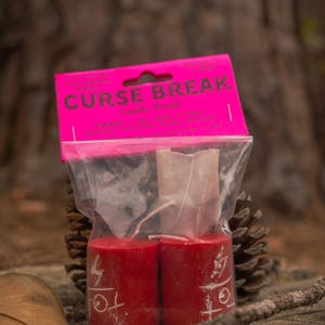 May include: A clear plastic bag containing two red cylindrical candles with white symbols printed on the front. The packaging features a bright pink header card with the text CURSE BREAK Candle Ritual printed in black capital letters.