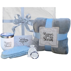 May include: A gift box set with a blue and gray theme. The set includes a gray throw blanket with the text "Stronger than the Storm", a white tumbler with the same text, a blue headband, a small white bag with a blue beaded bracelet, and a card that says "Thinking of you".
