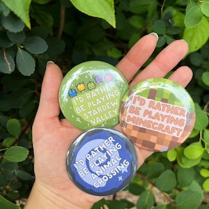 May include: Three round buttons with the text "I'D RATHER BE PLAYING" followed by "Stardew Valley", "Minecraft", and "Animal Crossing". The buttons are green, brown, and blue, held in a hand against a green leafy background.