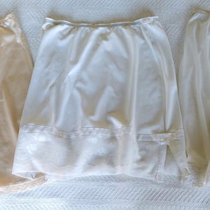 May include: Three vintage slips in the image. The slip on the left is a light peach color, while the middle and right slips are white. All three slips have a lace trim at the hem. The slip on the right is a pair of shorts.