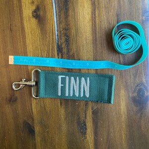 May include: A green fabric keychain with a silver clip and the name "FINN" embroidered in white.