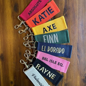 May include: A set of seven colorful keychains with different names embroidered on them. The keychains are made of fabric and have a silver metal key ring. The names on the keychains are: Absolutez, Katie, Axe, Finn, El Dorado, Bel Isle Eq., and Rayne.