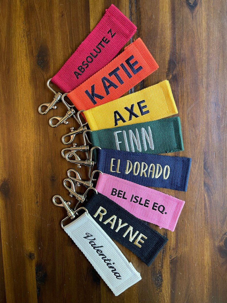 May include: A set of seven colorful keychains with different names embroidered on them. The keychains are made of fabric and have a silver metal key ring. The names on the keychains are: Absolutez, Katie, Axe, Finn, El Dorado, Bel Isle Eq., and Rayne.
