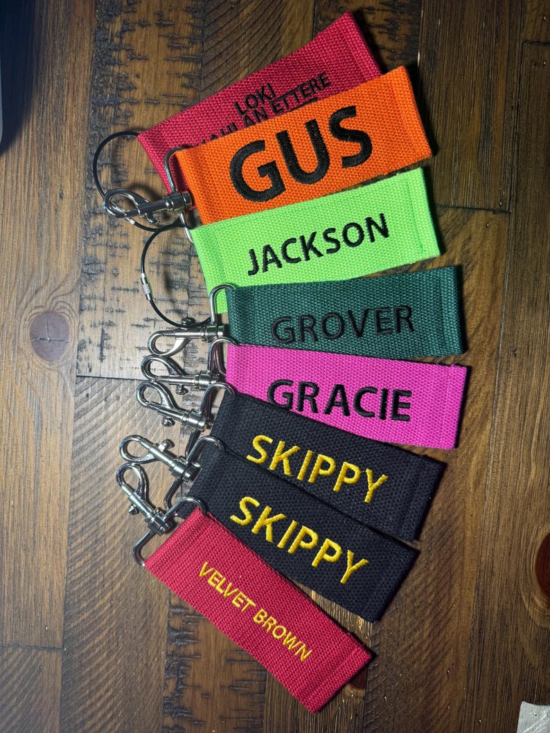 May include: A set of eight colorful keychains with different names embroidered on them. The keychains are made of fabric and have a silver metal ring. The names on the keychains are Gus, Jackson, Grover, Gracie, Skippy, Skippy, and Velvet Brown.