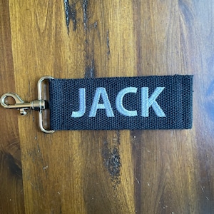 May include: Black fabric keychain with a silver metal ring and the name "JACK" embroidered in white.