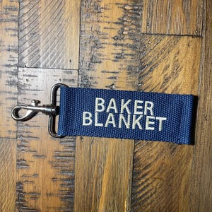 May include: A dark blue key chain with a silver metal clasp. The key chain has the words "BAKER BLANKET" embroidered in white.