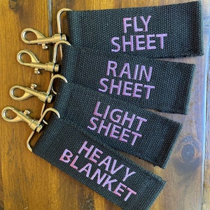May include: Four black fabric keychains with silver key rings. The keychains are labeled "Fly Sheet", "Rain Sheet", "Light Sheet", and "Heavy Blanket" in purple embroidery.